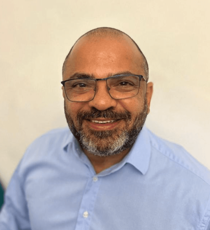 Homem careca sorridente com óculos e barba grisalha, vestindo uma camisa social azul clara.